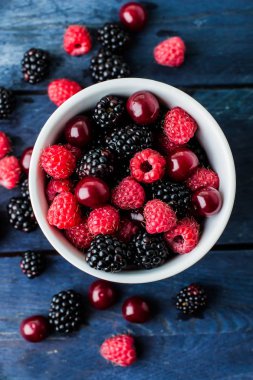 Mavi ahşap masa arka plan üzerinde Yaz taze çilek ile beyaz fincan üst görünümü. Kopya alanı. Vejetaryen gıda - ahududu, böğürtlen ve kiraz. Rustik stil