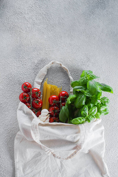 Reusable bag with groceries. Tote bag, minimal waste. Fresh basil, tomatoes cherry, garlic in fabric bag on dark table background. Top view, copy space.