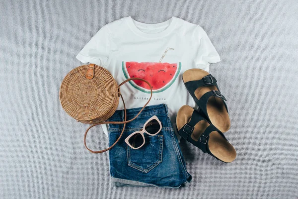 Flat lay with summer women\'s clothing and accessories. White t-shirt, blue denim shorts, fashionable organic rattan bag, sunglasses. Vacation, travel concept. Top view
