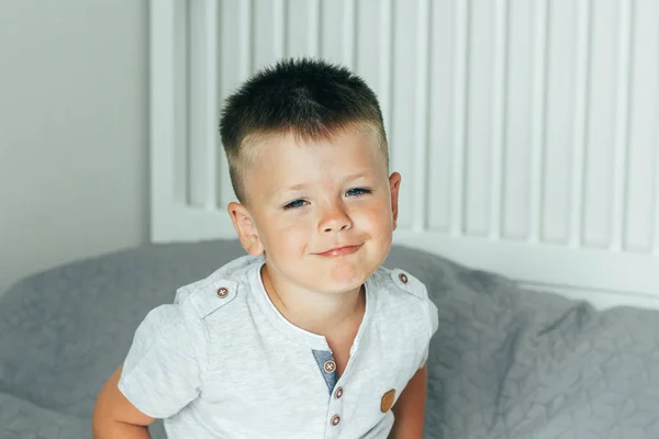 Portret 4 of 5 jaar oud beetje schattig glimlachend en plezier jongen op een bed in de slaapkamer. Een brunette jongen in een grijs overhemd. — Stockfoto