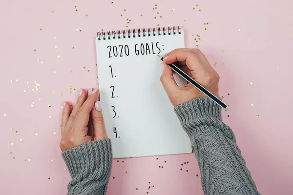 Feliz Año Nuevo 2020. Mano de mujer escribiendo 2020 Metas en cuaderno decoradas con decoraciones navideñas sobre fondo rosa. Vista superior, plano —  Fotos de Stock