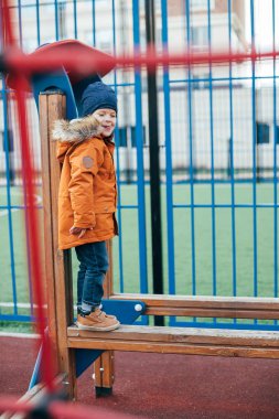 Çocuk 5 yaşında, çocuk parkında turuncu ceketli. Çocukların dışarıda boş zamanı, sokaklarda oyunlar..