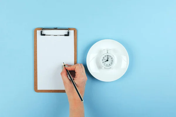 Intermittent fasting, ketogenic, diet concept. Female hand writting weight loss plan? Empty plate with white alarm clock on blue background. Flat lay, top view — Stock Photo, Image