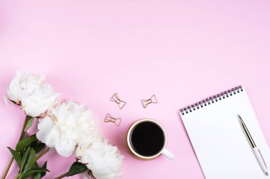 Kahve fincanı, boş defter ve pembe masa arkasına beyaz şakayık çiçekleri. Samimi, sakin bir hayat, asgari konsept. Üst görünüm, düz görünüm, kopyalama alanı
