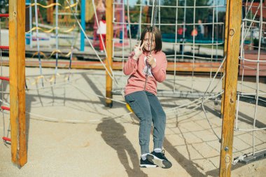 Şehir parkında oyun parkında 7-8 yaşlarında pembe kazaklı ve gri eşofmanlı tatlı beyaz bir kız. Açık hava aktiviteleri, yaşam tarzı, çocuk eğlencesi.