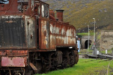Eski, paslı, terk edilmiş bir buharlı lokomotif kullanılmayan bir hat üzerine park edilmiş, önü manzaralı, dağı delip geçen bir tünel var.