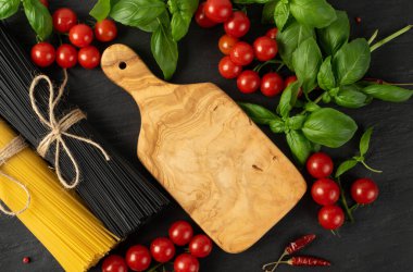Çiğ, ev yapımı makarna, zeytin ağacı kesme tahtası. Kuru siyah erişte, sarı makarna, kiraz domatesleri, yeşil fesleğen. Üst görünümü fotokopi alanı olan pişmemiş İtalyan spagtosu