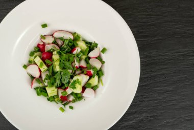 Bahar turpu salatasının üstü görünüyor. Siyah arka planda beyaz restoran tabağında taze salatalık ve yeşiller.