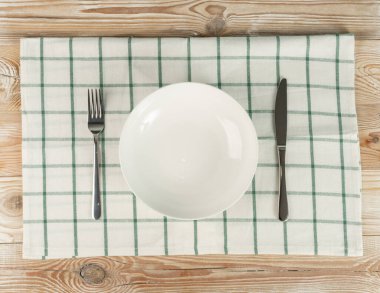 Boş beyaz tabak ve çatal bıçak takımı ahşap masa üzerinde fotokopi alanı ile birlikte. Kırsal mutfak kase, çatal ve bıçakla, kontrol edilen masa örtüsü veya bez peçeteyle modellenir.