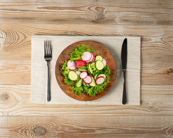 Spring homemade radish salad with fresh cucumber and greens on wooden plate top view. Simple green rustic salat with sliced radishes, cucumbers, escarole and lettuce