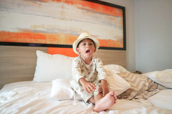Азиатский мальчик в львиной пижаме и соломенной шляпе сидит на кровати отеля с открытым ртом, глядя выразительно и весело