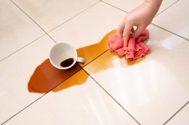 Dökülmüş kahve, kadın fayanslı zemine kahve lekesini siliyor, beyaz bardakla karışık sızıntı, kahverengi içki dökülmüş temizlik.