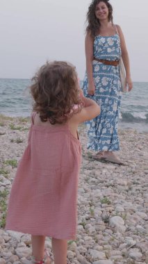 Küçük kız annesinin çakıl taşı plajında fotoğrafını çekiyor, deniz kenarında bir yaz tatilinin tadını çıkarıyor.