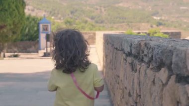 Genç bir kız turistik bir yerde, taş bir duvarın yanında yürüyor.