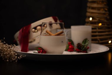 Tarçın çubuklarıyla süslenmiş bir bardak rompope, yanında Noel süslemeleri, sıcak ve şenlikli bir atmosfer yaratır.