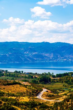 Ohri Gölü 'nün dolambaçlı yolu, yeşil tarlaları ve kabarık bulutlu parlak mavi gökyüzünün altındaki dağlarla manzarası