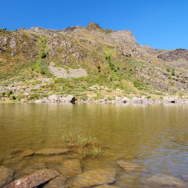 Berrak mavi bir gökyüzünün altında görkemli kayalık bir manzarayla çevrili sakin bir alp gölünün geniş bir görüntüsü..