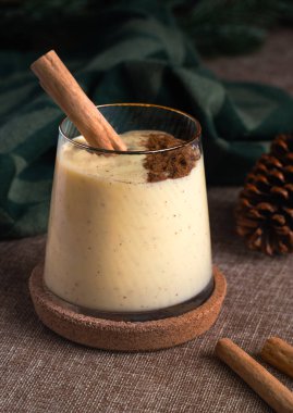 Bir bardak ev yapımı kremalı yumurta likörü, tarçın çubuğuyla süslenmiş ve fonda hindistan cevizi serpiştirilmiş, neşeli yeşil bir bez serpiştirilmiş.