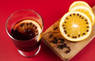 Üzerinde karanfil desenli portakal dilimi ile süslenmiş bir bardak sıcak şarap manzarası. Sahne kırmızı arka planda, iki portakal dilimi ve bol karanfil içeren ahşap bir kesme tahtasıyla sıcak ve davetkar bir kompozisyon yaratıyor.