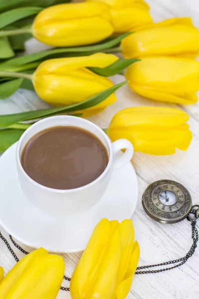 yellow tulips, a cup of coffee, cappuccino and vintage watch on a chain on a white wooden background