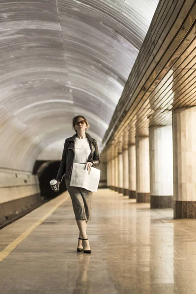 Business woman with a cup of coffee and a newspaper. Beautiful girl in a business suit and sunglasses with a cup of coffee and a newspaper at the subway station. A woman in a jacket. Success concept