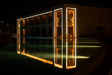 Lüks otelin aydınlanmış modern mimarisi geceleri huzurlu havuza yansıyor.