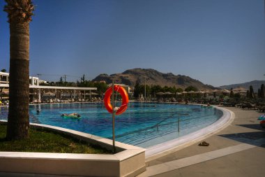 Lifebuoy on a stand near a bright blue swimming pool with mountains in the distance on a hot day