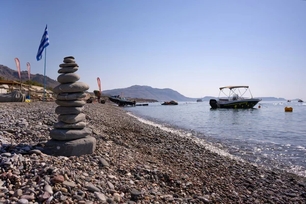 A balanced cairn made out of flat rounded rocks erected on a sunny pebble beach with flags and boats