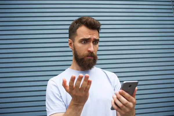 Handsome young man with a beard in a grey t-shirt looking puzzled, starring at mobile in his hand, the other hand is raised too on an urban setting background at daytime.