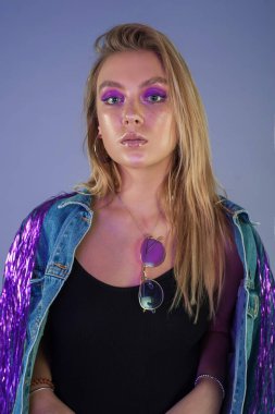 Uzun sarı saçlı ve etkileyici mor makyaj parlak saçak ile şık denim kıyafeti giymiş beyaz genç kız portresi izole Studio leylak arka plan üzerinde. Kameraya bakarak.