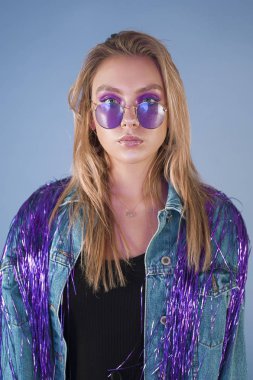Uzun sarı saçlı ve etkileyici mor makyaj mor güneş gözlüğü ve denim ceket Studio leylak arka plan üzerinde izole mor parlak saçak ile beyaz genç kız portresi. Kameraya bakarak.