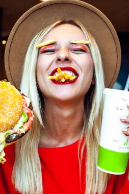 Güzel mutlu sarı saçlı Genç kız ve ağzı patates kızartması tam parlak yaratıcı makyaj gözleri fast food sahiptir kapa, holding burger ve her iki elinde içecek. Sağlıksız beslenme kavramı