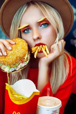 Güzel sarışın genç kız kırmızı ve ağzı fast food restoran çevrili patates kızartması tam oturan parlak yaratıcı makyaj bir şapka. Kameraya bakıyor. Sağlıksız beslenme kavramı