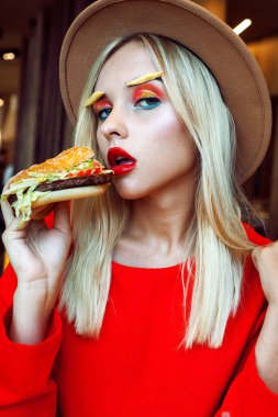 Genç güzel sarışın genç kız kırmızı parlak yaratıcı makyaj Restoran kameraya bakarak onun ağzında hamburger holding şapkası ile. Sağlıksız beslenme kavramı.