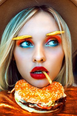 Güzel sarışın beyaz kız parlak yaratıcı makyaj ve kaşları yüzünü bir burger patates kızartması ağzına sahip dinlenme yerine patates ile portre portre. Sağlıksız beslenme kavramı.