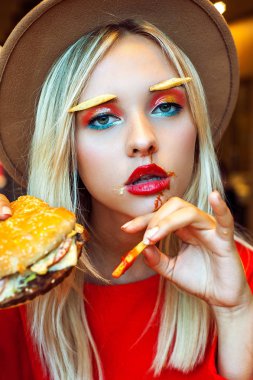 Güzel genç kız kırmızı ve parlak yaratıcı makyaj ve patates kızartması yerine restoranda yüzünde bir ketçap fast food yeme kaşları giyen bir şapka. Kameraya bakıyor. Sağlıksız beslenme kavramı.