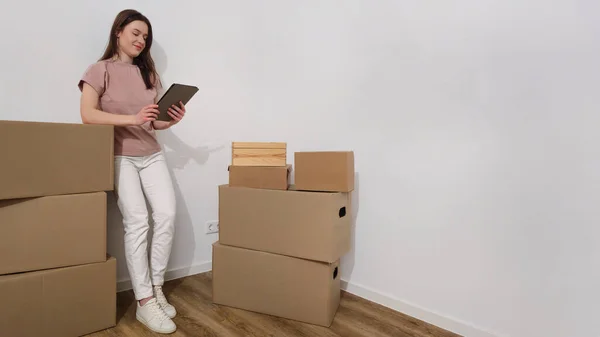 Young female student standing next to cardboard boxes, holding a tablet in a bright room with wooden flooring, illustrating student moving process and home organization.