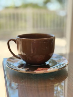 Çiçekli çay tabağının üzerinde çay ve yansımayı bekleyen boş kahverengi bir çay fincanı. Parlak bir günde cam bir masanın üzerindeki parlak kahverengi seramik..