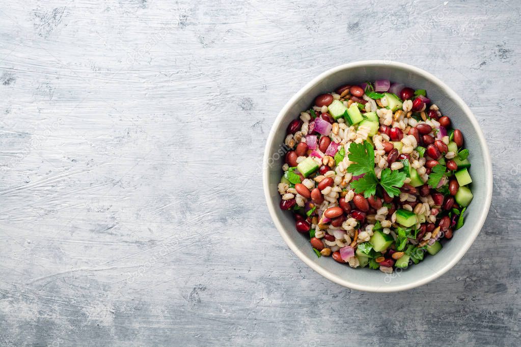 Healthy pearl barley salad with beans, cucumbers, red onion, sunflower seeds, pomegranate and parsley in bowl on concrete background. Top view. Copy space.