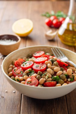 Ahşap masada kase domates, kapanlar, sarımsak ve maydanoz ile beyaz fasulye salatası. Seçici odak.