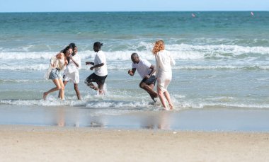 Group of diverse friends having fun at the beach, running and splashing in the waves on a sunny day