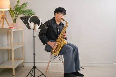 Kapalı bir odada, yumuşak ışıklandırmalı saksafon çalan genç bir Asyalı. Ev müziği stüdyosunda rahat bir solo performans.