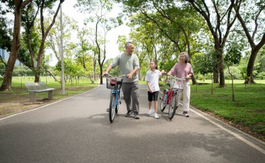 Mutlu Asyalı büyükanne ve büyükbaba bisikletleriyle parkta torunlarının yanında yürüyor, aile açık havada güzel vakit geçiriyor, huzurlu bir günde konuşuyor ve gülümsüyor.