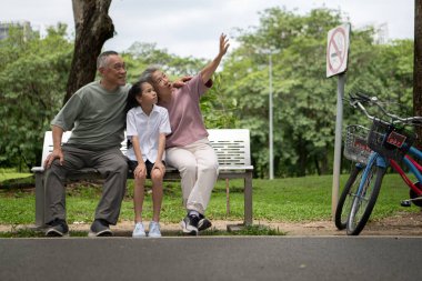Mutlu Asyalı büyükanne ve büyükbaba bisikletleriyle parkta torunlarının yanında yürüyor, aile açık havada güzel vakit geçiriyor, huzurlu bir günde konuşuyor ve gülümsüyor.