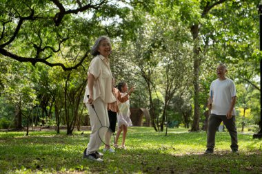 Mutlu Asyalı büyükanne ve büyükbaba torunlarıyla yeşil bir parkta piknik yaparken güneşli bir günde gülüp kaynaşırken torunları badminton oynuyorlardı.,