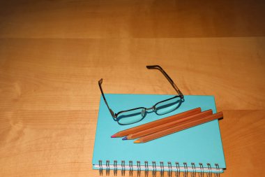 Ahşap bir masanın üzerinde zarif okuma gözlüğü ve üç renkli kalemle kaplı mavi sarmal bir defter bulunur. Eğitimi, eğitimi ve yaratıcı düşünceyi sembolize eden yaratıcı bir kompozisyon..