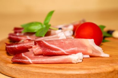 Narin prosciutto dilimleri, olgun, sulu domatesler, mis kokulu fesleğenler ve tüm siyah karabiberlerle çevrili, ahşap bir tahta üzerinde ustalıkla düzenlenmiş.. 