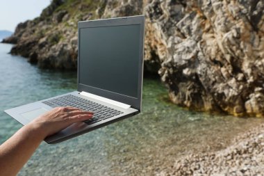 Kaya kıyıları yakınında dijital göçebe yaşam tarzını ve özgürlüğünü simgeleyen bir dizüstü bilgisayar kullanan bir kişi olarak deniz kenarında uzaktan çalışma.