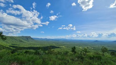 Etkileyici bir panoramik manzara dağların enginliğini ve açık mavi gökyüzünün altında, tüylü beyaz bulutlarla benekli yeşil bir vadiyi yakalıyor. Geniş yağmur ormanları, güneş doğarken zengin biyolojik çeşitlilik sergiliyor, dingin ve huzurlu bir atmosp yaratıyor.