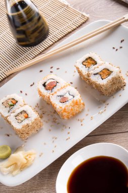 Sushi on a white square plate with soy sauce and natural wooden chopsticks on a light wooden table.